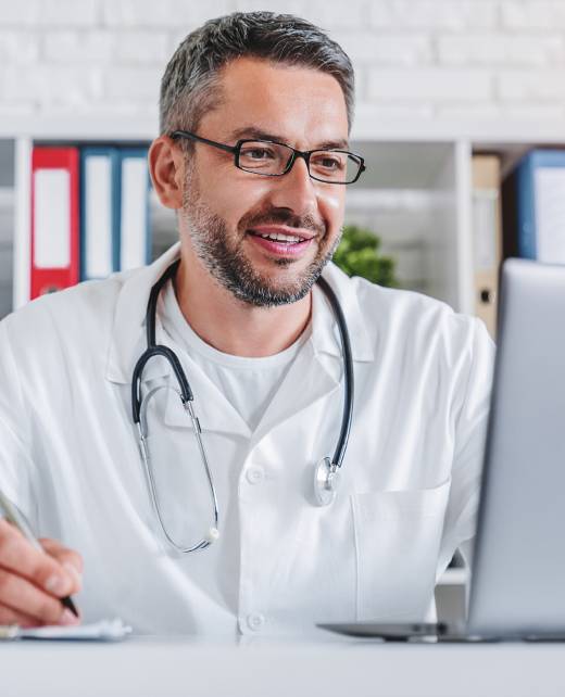 Adult male doctor working on laptop writing prescription sits at desk.